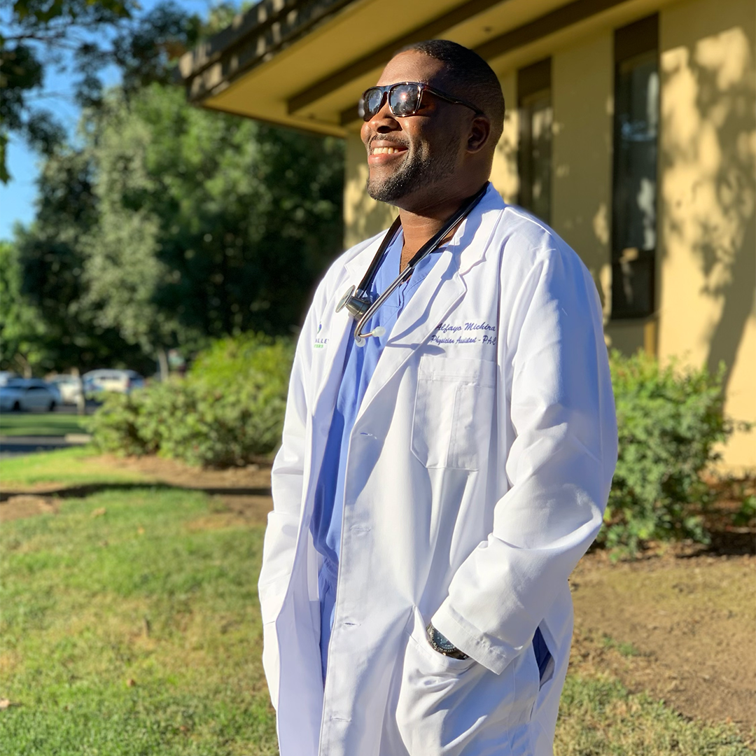 Alfayo Michira stands outside in PA white coat and sunglasses, smiling
